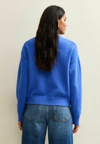 Blauwe gebreide trui met een relaxte pasvorm en ribbelzoom, gecombineerd met hoge taille blauwe jeans, gezien vanuit de achterkant.