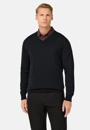 Schwarzer V-Ausschnitt-Pullover aus weichem Strickstoff, mit langen Ärmeln und gerippten Bündchen. Getragen über einem dunkelbraunen Kragenhemd.