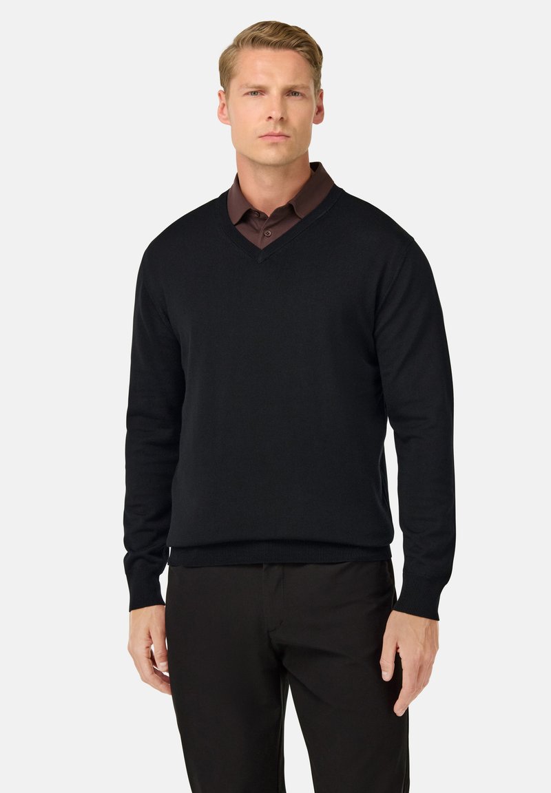 Sweater noir à col en V fabriqué en tissu tricoté doux, avec des manches longues et des poignets côtelés. Porté par-dessus une chemise à col marron foncé.