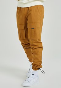 Cargo-byxor i senapsgul tyg med en texturerad yta, utrustade med stora cargo-fickor och elastiska ankle cuffs. Bärs med vita sneakers.