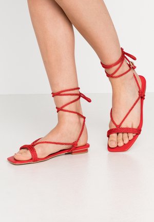 Czerwone płaskie sandały z kwadratowym noskiem, z plecionym wzorem sznurkowym oplatającym kostkę. Gładkie matowe wykończenie i niski obcas.