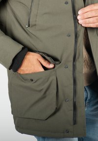 Veste extérieure verte avec fermeture éclair sur le devant, grandes poches avant et poignets côtelés. Le matériau semble lisse et résistant à l'eau.