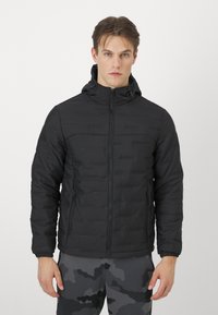 Giacca a vento nera con zip frontale, cappuccio e polsini elastici, caratterizzata da un design a quadri rettangolari. Indossata sopra pantaloni grigi con motivo camouflage.