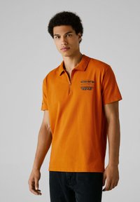 Oranžová polo tričko so zipsovým golierom, s modrým grafickým textom a logom na prednej strane, vyrobené z hladkého bavlneného materiálu.