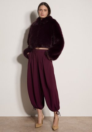 Femme portant une veste courte en fourrure violet foncé, un pantalon violet ample taille haute resserré aux chevilles, et des chaussures à talons beiges.
