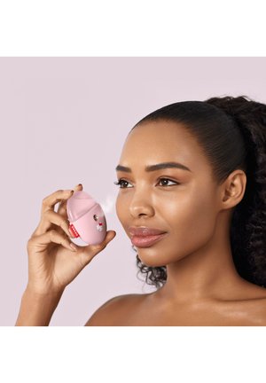 Kvinde med krøllet hår bruger en lyserød ansigtsmist-enhed, der udsender spray nær hendes kind mod en lys lilla baggrund.