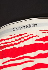 Svart tyg med Calvin Klein-logga på en elastisk midjeband; den nedre delen har ett mönster av röda vågiga linjer på en ljus bakgrund.