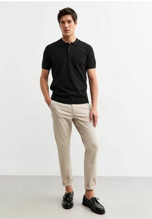 Polo shirt nero a maniche corte con colletto e tre bottoni, abbinato a chinos beige chiaro arrotolati e scarpe nere con lacci.