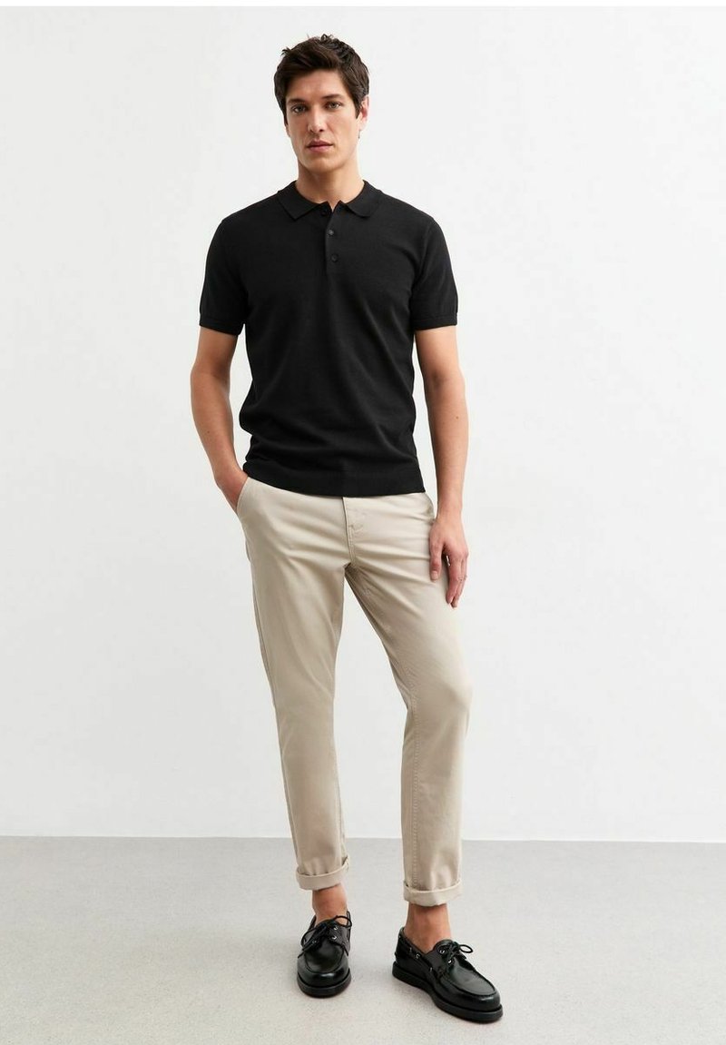 Zwarte korte mouwen polo met een kraag en drie knopen, gecombineerd met lichtbeige opgerolde chino's en zwarte veterschoenen.
