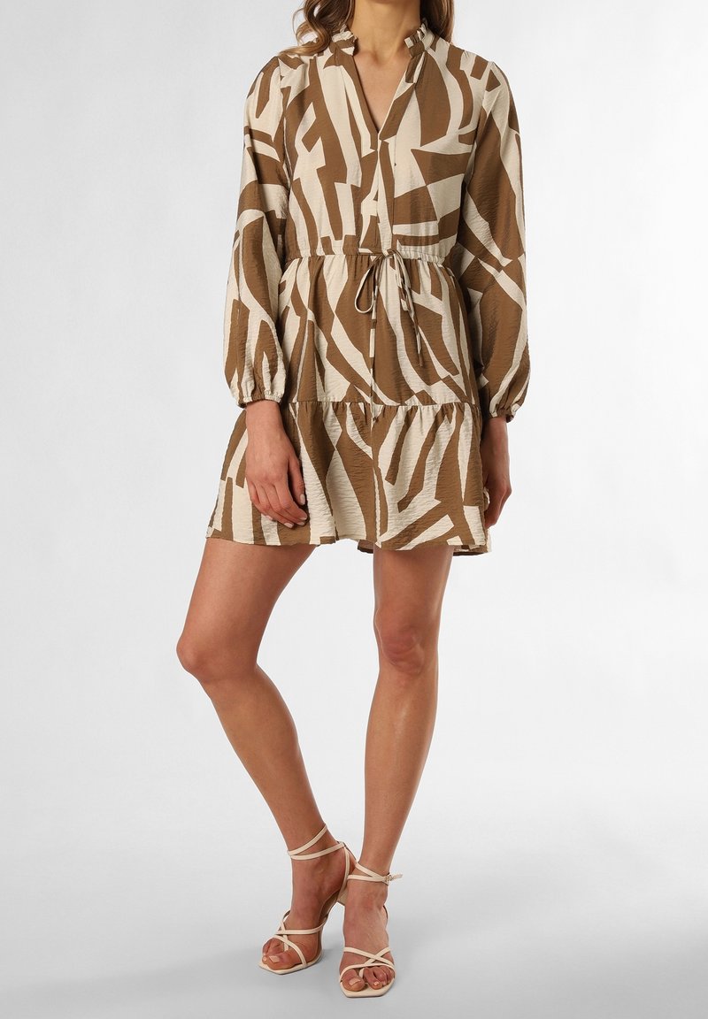 Femme portant une robe courte à motif abstrait beige et blanc avec manches longues et talons beiges à brides, debout devant un fond uni.