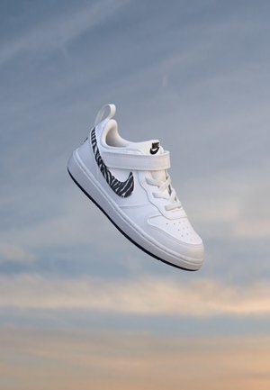 Baskets blanches Nike avec logo rayé zébré noir et fermeture velcro flottant sur un fond de ciel bleu et orange doux.