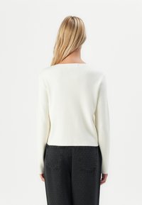 Blonde vrouw die met haar rug naar de camera staat, gekleed in een lange mouw witte trui en gestructureerde zwarte broek met achterzakken, tegen een effen achtergrond.