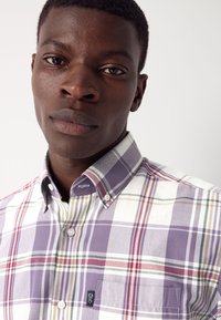 Mees, kes kannab kinnitatud ruudulist särk, millel on lillad, punased ja rohelised triibud, rinnatasku ja nupukraega, lähivaade.