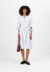 Mulher em pé, vestindo um vestido midi listrado em branco e azul com mangas atadas, segurando uma mala bordô e usando sapatos baixos de trama preta.