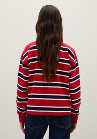 Rode lange mouwen shirt met witte en marineblauwe horizontale strepen, ronde halslijn en ribgebreide manchetten. Draagt met donkerblauwe jeans, achteraanzicht.