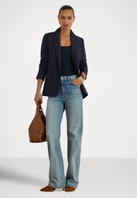 Moteris dėvi tamsiai mėlyną švarką su dryželiais, juodą palaidinę, šviesiai mėlynas džinsus, rudus smailius batelius ir laiko rudą rankinę, viena ranka kišenėje.