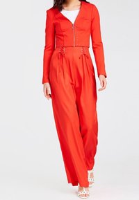 Rode jumpsuit met lange mouwen, een rits aan de voorkant en vetersluiting accenten bij de taille; gemaakt van gladde stof met brede pijpen.