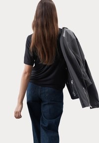 Zwarte t-shirt, relaxte pasvorm, gecombineerd met donkerblauwe wijde jeans, matte zwarte leren jas over de schouder, haar van gemiddelde lengte in een donkere kleur.