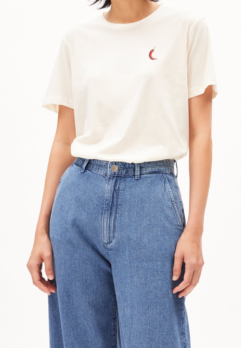 Beige bomullströja med kort ärm och en liten broderad röd chilifrukt på vänster bröst, i kombination med ljusblå denimbyxor.