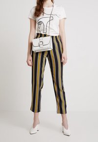 Vit t-shirt med en svart linjetegning, svart och gul randiga byxor, vit handväska med nitar, och vita spetsiga skor.