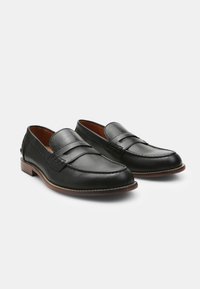 Mocasines de piel negra con un pequeño diseño recortado, detalles de costura y una suela marrón claro. Presentan una forma clásica de punta redondeada.