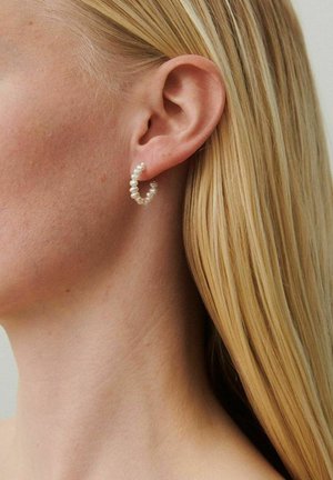 Plavolaska ženska, ki nosi majhno uhan v obliki obročka iz belih biserov, prikazuje svoje levo uho in stran vratu.