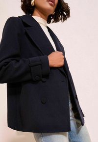 Femme portant un manteau bleu marine à double boutonnage avec de grands boutons, sur un haut blanc et un jeans bleu clair, sur un fond clair.