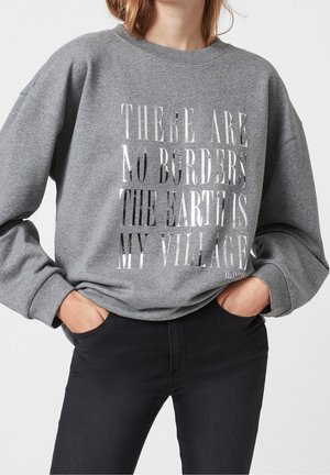 Persona con un sudadera gris con texto reflectante "No hay fronteras, la tierra es mi aldea" y pantalones negros, con las manos en los bolsillos.