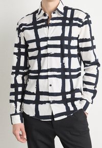 Chemise blanche avec motif à carreaux abstraits bleu marine, manches longues, boutonnée à l'avant, col pointu. Matière légère, texture lisse.