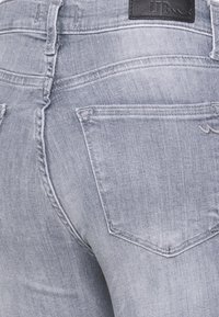 Calças de ganga azul claro com um corte slim, apresentando dois bolsos traseiros e detalhes de costura discretos. Logótipo da marca visível na cintura.