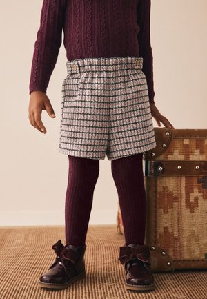 Kind draagt een bordeauxrode kabelgebreide trui en panty's, grijze geruite korte broek en glanzende bordeauxrode laarzen met strikjes, staand naast een patroonrijke koffer.