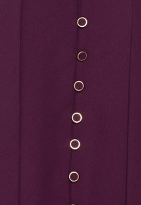 Rząd sześciu złotych okrągłych guzików na teksturowanej głębokofioletowej tkaninie, równomiernie rozmieszczonych w pionowej linii.