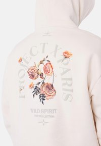 Sudadera crema con rosas bordadas en tonos rosa y naranja. El texto incluye "PROJECT X PARIS" y "WILD SPIRIT PXP COLLECTION" en gris.