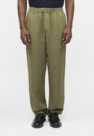 Άνδρας με ελαφρώς φαρδιά χακί παντελόνια με ελαστική μέση και μαύρα δερμάτινα παπούτσια, στέκεται μπροστά σε απλό φωτεινό φόντο.