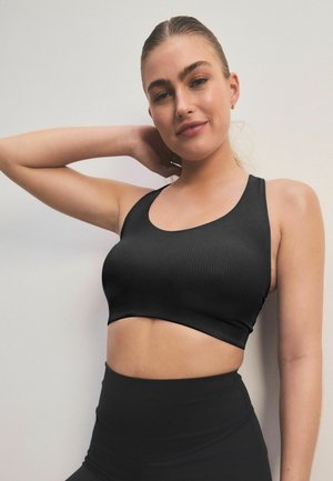 Soutien-gorge de sport noir côtelé avec un décolleté arrondi et des bretelles larges, associé à un legging noir taille haute, sur fond uni.