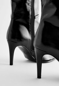 Juodos odinės kulkšninių batai su aukštu stiletu kulnu, turinčiu šoninius užtrauktukus ir lygią tekstūrą, demonstruojantys elegantišką dizainą.
