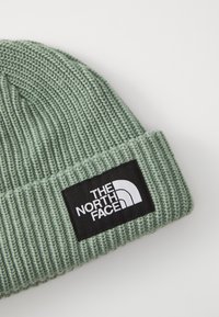 Bonnet en tricot vert avec une texture côtelée et un revers replié, présentant un patch logo rectangulaire noir avec du texte blanc et un design incurvé.