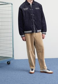 Giacca in corda blu navy con dettagli bianchi e ricami, abbinata a pantaloni in corda beige e sneakers bianche con lacci blu navy e suole marroni.