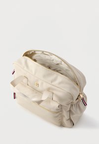 Beżowa torba z nylonu z złotym wykończeniem, dwoma uchwytami i dużym zapięciem na zamek. Posiada przednią kieszeń na zamek oraz kontrastowe akcenty pasków.