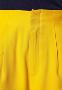 Gros plan sur un pantalon jaune avec des plis et une fermeture devant dissimulée, porté avec un haut bleu foncé.