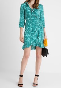 Zelenomodrá zavinovací šaty s bílými puntíky, volánkovým výstřihem, tříčtvrtečními rukávy a rozšířeným lemem. Doplněno černými páskovými lodičkami.