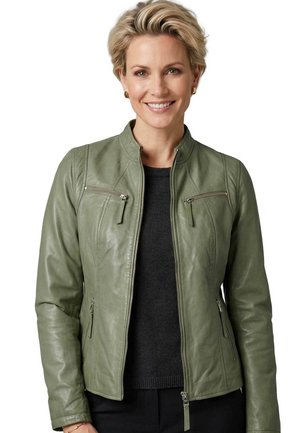 Lächelnde Frau mit kurzen blonden Haaren, die goldene Ohrringe, eine grüne Lederjacke mit Reißverschluss und einen schwarzen Pullover vor weißem Hintergrund trägt.