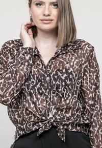 Chemise imprimée animale marron et crème avec un devant noué, design à boutons, manches longues et col classique. Tissu léger et texturé.