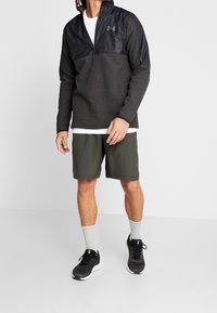 Hombre con un pulóver de media cremallera en gris oscuro y negro, pantalones cortos oliva, calcetines blancos y zapatillas negras de Under Armour, de pie contra un fondo blanco.