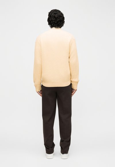 Άτομο με σγουρά σκούρα μαλλιά στέκεται με την πλάτη, φοράει εκρού πλεκτό πουλόβερ, σκούρο παντελόνι και λευκά αθλητικά παπούτσια σε απλό φόντο.