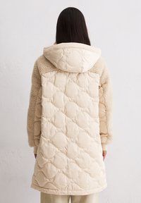 Frau mit langen dunklen Haaren, die einen knielangen beigen gestepp-ten Mantel mit Kapuze und FleecefÃ¤rmel trÃ¤gt, die an einer weiÃen Wand steht.