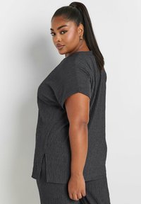 Musta teksturoitu yläosa lyhyillä hihoilla, jossa on rento istuvuus ja sivuhalkiot liikkumisen helpottamiseksi. Sujuva, rypistynyt kangas.