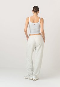 Kobieta stoi tyłem, ubrana w biały top na ramiączkach, jasnoszare dresy, białe trampki, a jej włosy są związane w kok, na jednolitym tle.