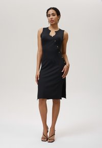 Schwarzes geripptes Kleid mit einem Schlüssellochausschnitt, goldenen Knöpfen an der Seite und einem schmalen Schlitz bis zum Oberschenkel. Der Stoff ist glatt und hat eine figurbetonte Form.