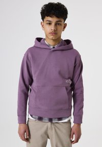 Chico adolescente vistiendo una sudadera morada sobre una camisa de cuadros y pantalones beige, de pie frente a un fondo blanco liso, mirando hacia adelante.